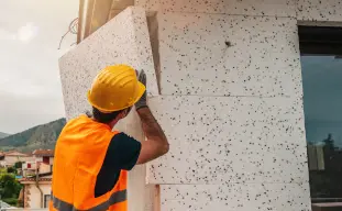 Homme portant un gilet orange et un casque de chatier blanc qui installe des blocs de polystyrene blanc sur un mur d'une maison