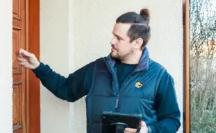 homme portant une doudoune sans manche Tucoenergie qui frappe à la porte d'une maison et qui tient une tablette noire