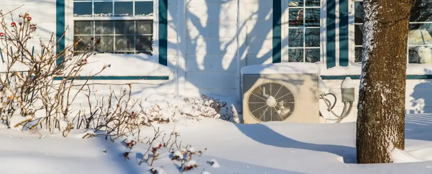 une pompe à chaleur sous la neige devant une maison en hiver